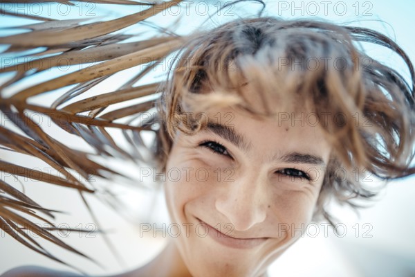 A joyful teenage boy smiles under the dappled shade of a palm tree, his hair tousled by a gentle breeze. The bright blue sky in the background enhances his cheerful demeanor