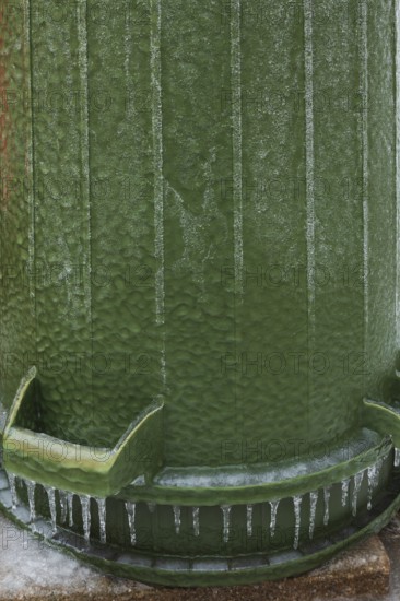 Upside down green PVC and rubber thrash can covered with thick sheet of ice and hanging icicles after freezing rain in late winter, Quebec, Canada
