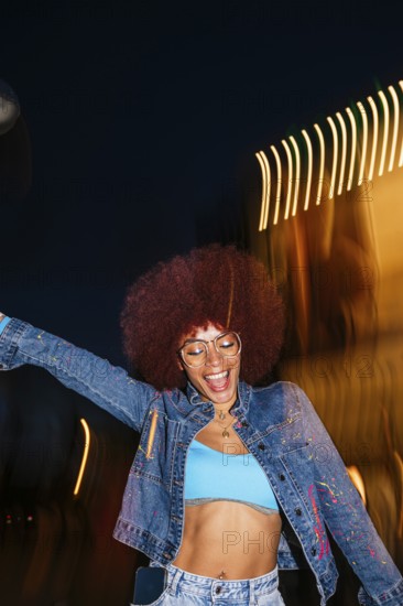 African descendant woman with vibrant afro hair joyfully dancing under night lights. Wearing glasses and a denim jacket, she embodies a lively urban scene with eyes closed