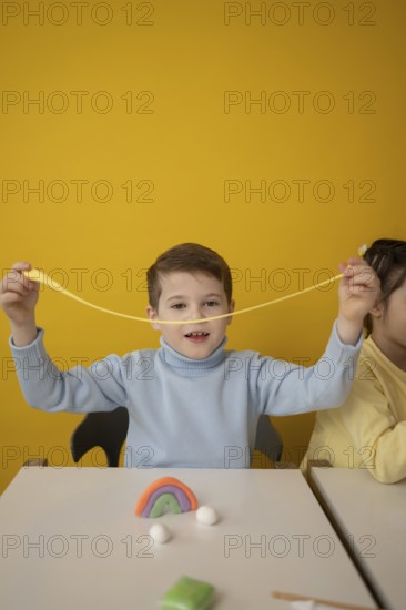 A child joyfully stretches colorful modeling clay during a creative lesson. The bright yellow background enhances the playful atmosphere, promoting imagination and expression