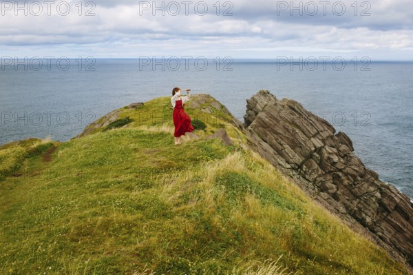 A woman clad in a flowing red dress stands on the rugged cliffs of Middle Cove overlooking the Atlantic Ocean, Newfoundland & Labrador. The image captures a serene moment against a scenic natural backdrop