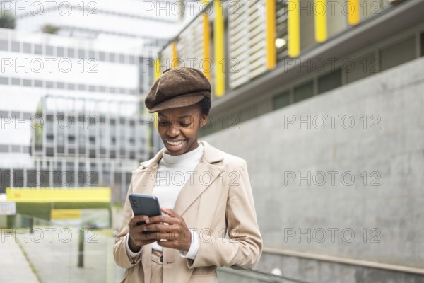 An urban black woman smiles while checking her phone in the city She wears a beige coat and cap, embodying a modern and stylish look The scene captures urban elegance and connectivity