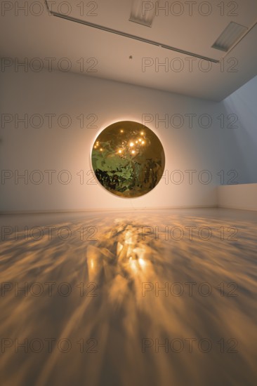 A round mirror on the wall casts warm light reflections onto the floor in an art space, Schauwerk Sindelfingen, Germany