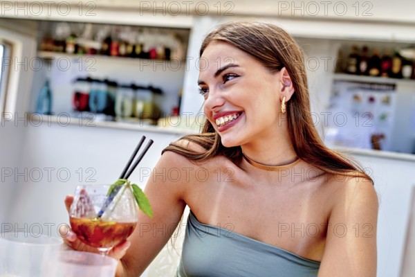 A woman smiles while holding a colorful cocktail adorned with mint at a lively beach bar. The atmosphere is relaxed, perfect for summer enjoyment with friends