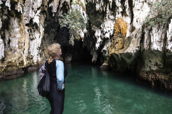 A woman stands at the edge of a cave, admiring the striking rock formations and vibrant turquoise water in Indonesia. The scene captures the essence of discovery and adventure