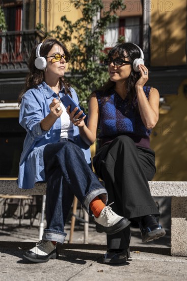 A multiethnic lesbian couple sits on a bench outdoors, wearing headphones and sharing a joyful moment while enjoying music under the sunlight, symbolizing unity and pride