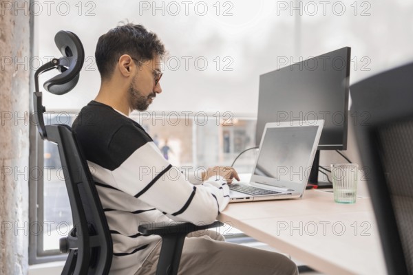 A professional concentrates on his laptop in a modern office environment Natural light illuminates the workspace, emphasizing focus and productivity in a collaborative setting