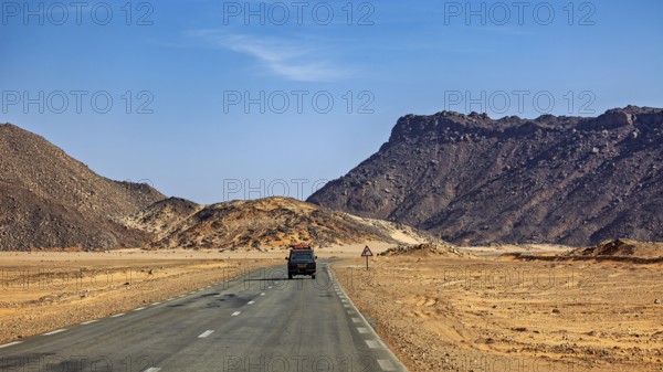 An off-road vehicle drives on a tarmac road through a mountainous desert landscape, With an off-road vehicle through the endless landscape of the Sahara desert in Algeria