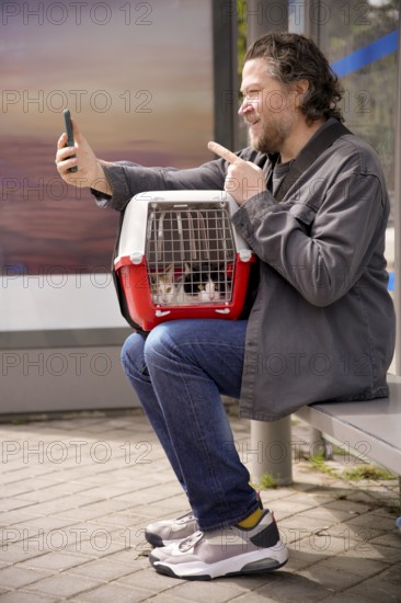 A man sits at a bus stop with two cats in a carrier on his lap, taking a selfie with his smartphone. The scene captures a light-hearted moment in urban life