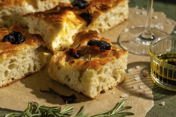 Golden homemade focaccia topped with sun dried tomato and rosemary. Served with olive oil, this savory delight rests on parchment paper, offering a rustic and appetizing look