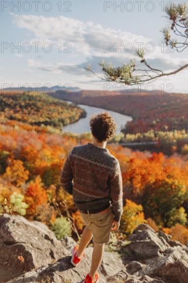 A person stands on a rocky overlook, admiring the vibrant autumn foliage in Quebec, Canada. The river winds through the forest, surrounded by colorful trees under a clear sky