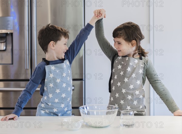 Two young siblings in star patterned aprons enthusiastically raise their hands in celebration while baking in a modern kitchen, capturing joyful teamwork and family bonding