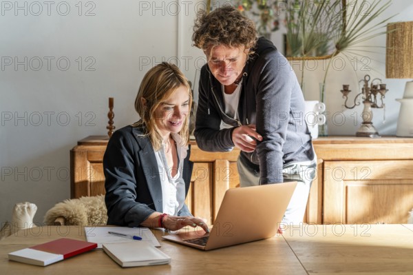 A couple works together at a home office, focused on a laptop The room is cozy, with natural lighting Papers and pens are on the table, emphasizing productivity