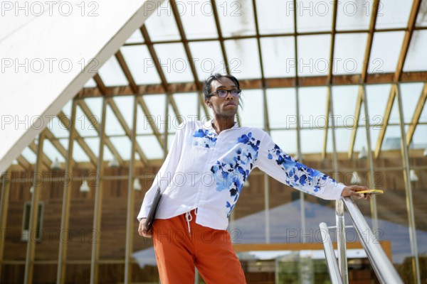 A man in a vibrant, floral shirt and orange pants poses confidently near a modern glass building, expressing pride and individuality. Represents fashion and diversity