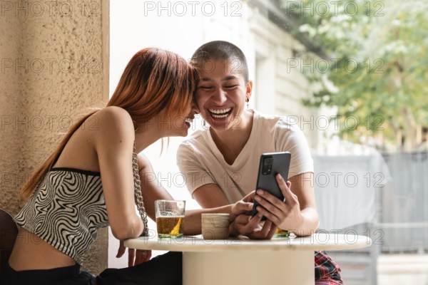 Two lesbian women share a joyful moment at a cafe, laughing and looking at a smartphone. They sit at an outdoor table with drinks, surrounded by a warm and inviting atmosphere