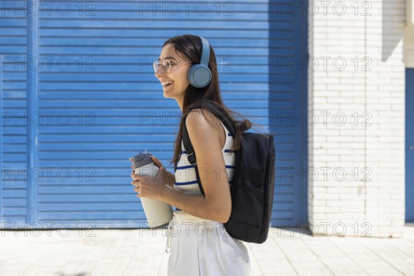 A woman enjoys city life outdoors, wearing headphones and carrying a water bottle, embodying an active and modern lifestyle. She is dressed casually with a backpack, reflecting urban chic