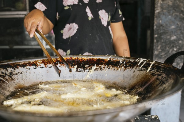 A woman prepares street food in a large wok using tongs. The image captures the sizzling oil and the dynamic culinary process in an outdoor setting in Bali