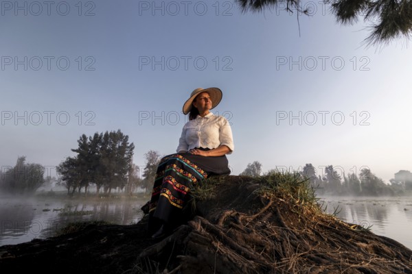 A woman in traditional attire enjoys a peaceful moment by the water in Xochimilco, Mexico. Surrounded by nature, she reflects the region's rich cultural heritage and tranquil beauty