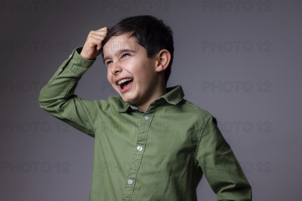 A young boy in a green shirt is laughing joyfully against a gray background. His expressive eyes and bright smile capture a moment of carefree happiness and childhood innocence