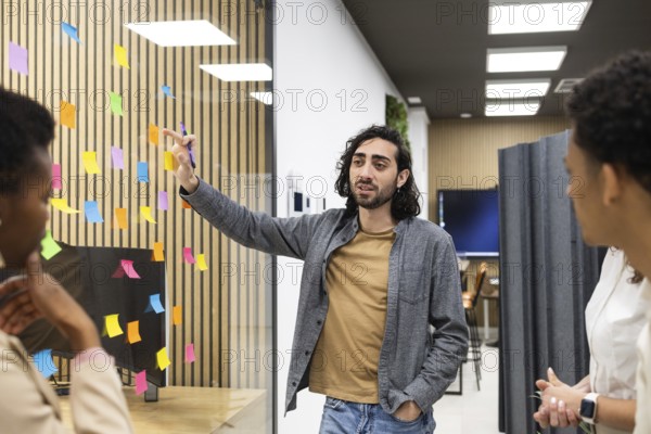 Professionals engage in a brainstorming session in a modern office space. A man gestures towards colorful sticky notes on glass, fostering creativity and collaboration