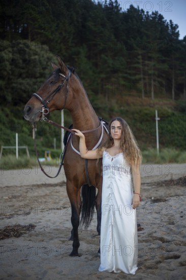A young woman in a flowing white dress stands beside her horse on a tranquil beach. The scene captures a blend of elegance and natural beauty, with a serene forest backdrop