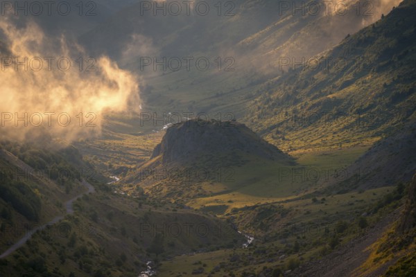 A stunning valley landscape at sunrise, with golden light illuminating mist and rugged hills in the Pyrenees. Shadows and highlights create a serene and majestic atmosphere in summer