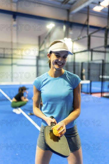Young woman smiling and holding a pickleball paddle with a perforated ball, standing on a bright blue indoor court, ready for active sport and recreational play