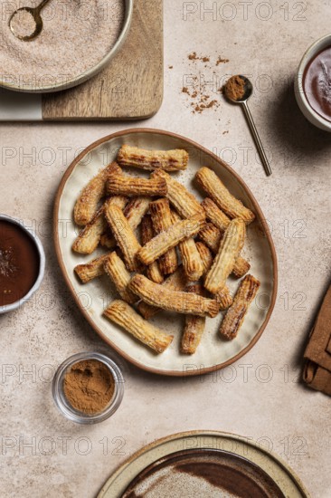 Top view of a ceramic plate filled with sugary churros next to bowls of chocolate sauce and cinnamon, set against a background of rustic linen and wooden kitchenware