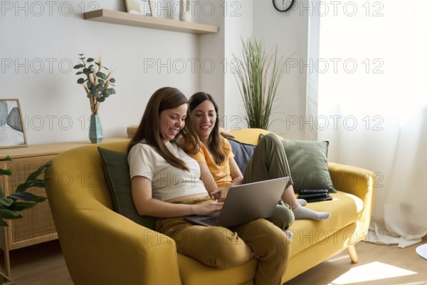 A lesbian couple sits comfortably on a yellow couch, smiling and using a laptop together in a cozy living room, highlighting love and togetherness in the LGBTQIA+ community