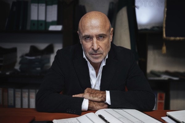 Portrait of a confident businessman seated at a desk, surrounded by office files. The scene conveys focus and determination in a professional corporate setting