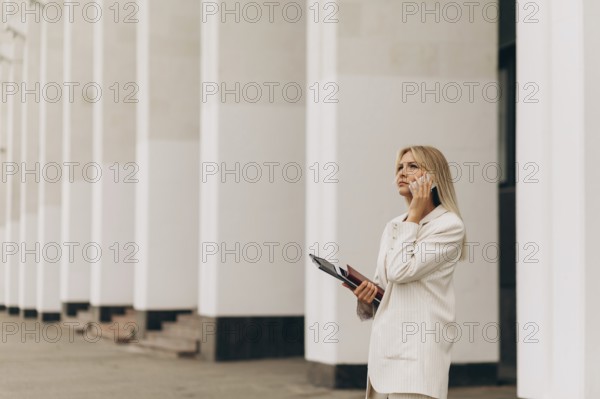 A businesswoman in a beige suit talks on her phone while standing outside an office building with white columns. She holds a folder, looking focused and professional