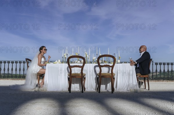 A picturesque wedding scene unfolds in an ancient villa in Treia, Macerata. The bride and groom are seated at an elegantly decorated outdoor table under a clear blue sky