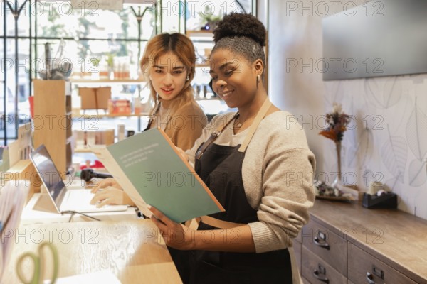 Two workers in a cosmetics store engage in tasks at the counter. One reads a catalog while the other types on a laptop. The store is bright, filled with beauty products