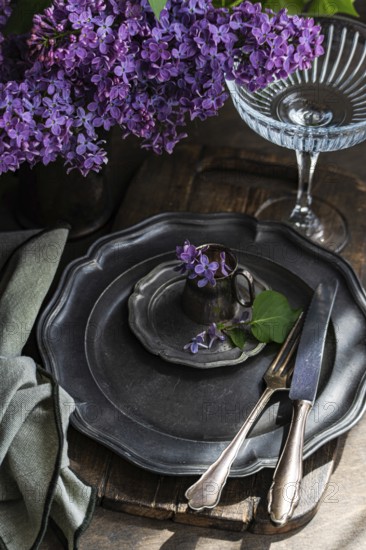 A rustic table setting features antique pewter plates and cutlery, adorned with vibrant lilac flowers in a vintage cup. A pleated napkin and glass add elegance to the scene