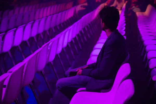 A indian man on a white chair in a spacious, purple lit auditorium at a conference of AI in Amsterdam. The atmosphere is serene with rows of empty seats