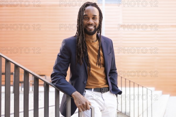 Cuban business man with long hair wearing a blazer and casual attire, leaning against a staircase railing outdoors, smiling warmly. Stylish urban setting with orange wall