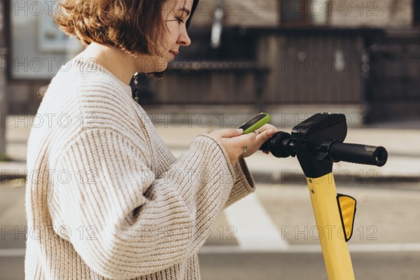 A woman is engaged in scanning a QR code with her smartphone to rent an electric scooter, standing outdoors on a sunny day. The scene captures her as she focuses on her mobile device, enabling urban mobility with modern technology