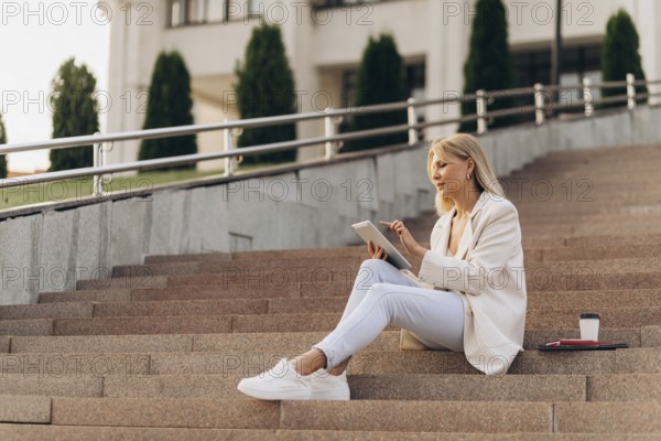 A businesswoman in casual attire sits on outdoor steps with a digital tablet, concentrating on her work. A takeaway coffee cup and papers lie beside her, emphasizing a modern urban lifestyle
