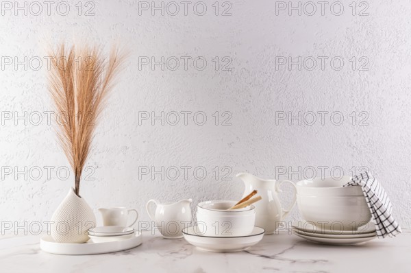 A minimalist interior design featuring elegant white tableware and a decorative vase with dried grass, arranged tastefully on a marble surface against a textured white wall