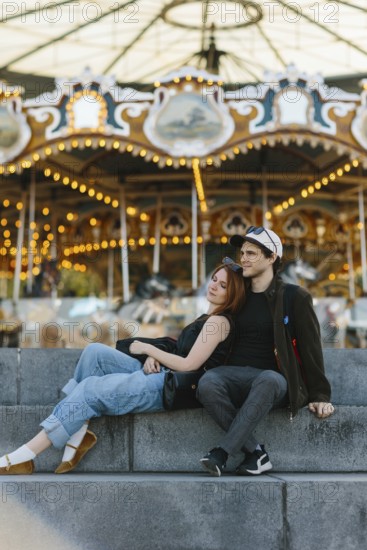 A couple sits on stone steps, the woman leaning on the man, in front of a brightly lit carousel in Brooklyn Bridge Park. They exude a sense of relaxation and enjoyment