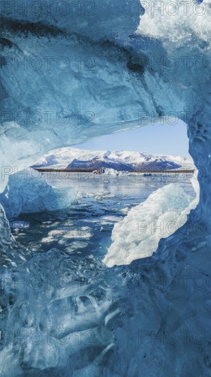 Stunning view from within a crystal-blue ice cave in Jokulsarlon, Iceland, framing the glacier lagoon and floating icebergs with snow-capped mountains in the background