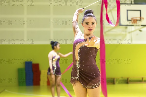 Young disabled rhythmic gymnast in elegant attire performs with a pink ribbon inside a sports hall. Another gymnast practices in the background. Dynamic movement and focus