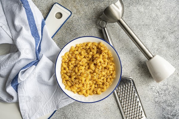 A bowl of chifferi rigati pasta sits on a counter top, accompanied by a grater, a mixer, and a dish towel. The scene captures a rustic kitchen atmosphere