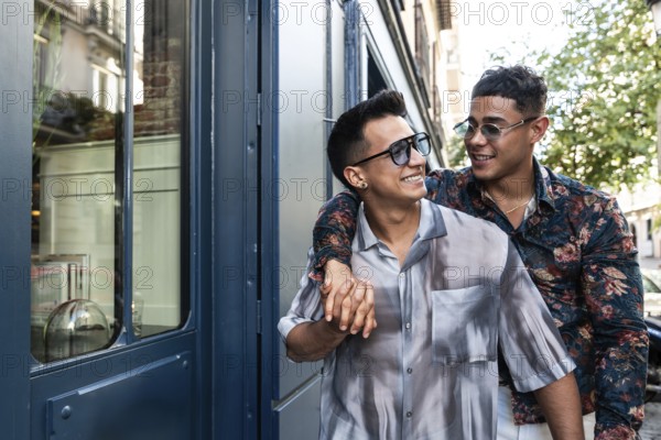 A cheerful latin gay couple walks hand in hand on a sunny day in Madrid. They share smiles and laughter, surrounded by the city's urban charm and lively atmosphere