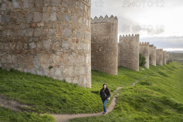 An Asian female tourist strolls along Avila's ancient medieval walls. The scene captures cultural exploration and architectural beauty