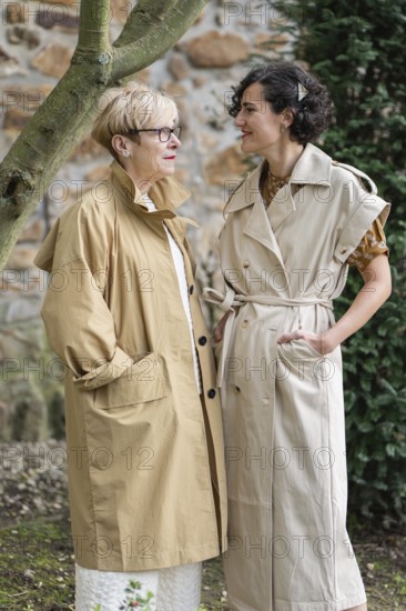 Two fashion women in stylish beige trench coats share a joyful moment outdoors. They stand near a stone wall and a tree, enjoying each other company in natural surroundings
