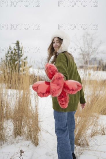 A woman wearing a green sweater and fur hat holds red heart shaped balloons in a snowy park. She smiles over her shoulder, embracing the winter scenery