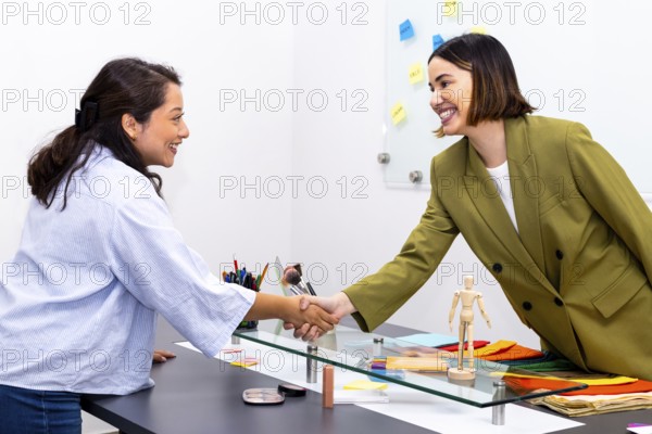 A vibrant image of two women shaking hands in an image consulting setting. They smile warmly, embodying professionalism and collaboration, with colorful fabric swatches on display
