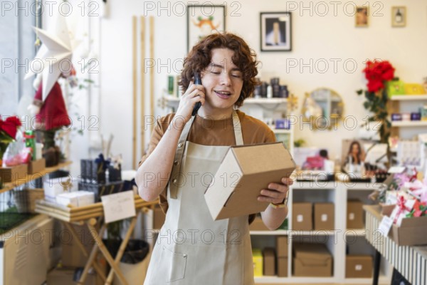 A young shop assistant wearing an apron talks on the phone while holding a cardboard box in an organic products store The shop's eco-friendly items and decor create a modern, natural vibe
