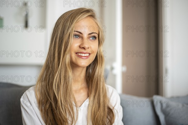 Portrait of a smiling woman with long blonde hair sitting in a bright, cozy room. She looks relaxed and content, capturing a moment of genuine happiness and warmth